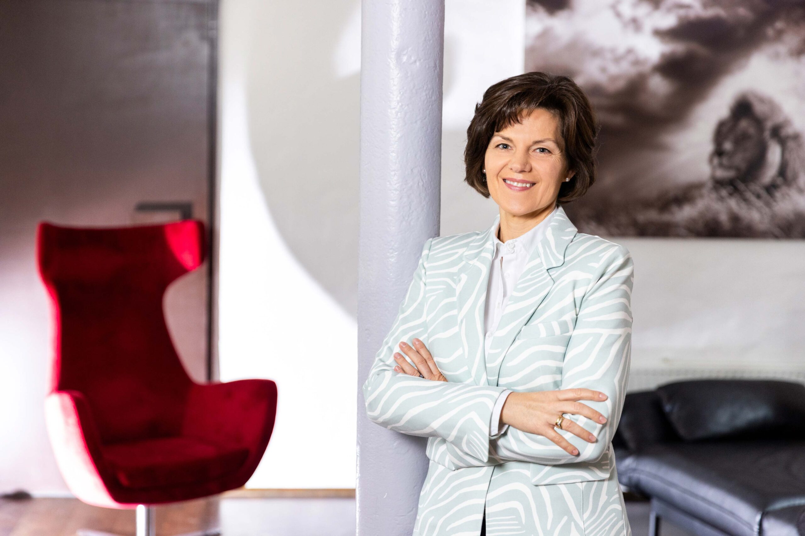 Katrin Pingel professionelle Coachin für Finanzen und Vorsorge aus Unna. Frau Pingel steht im Halbprofil mit verschränkten Armen an einer Säule angelehnt. Sie trägt einen Blaser mit Zebra-Muster in einem gellen Blau-Grau. Sie schaut lächelnd, sympathisch und einladend in die Kamera. Im Hintergrund steht ein roter Ohrensessel. Das Bild dient zur Illustration der Kontaktseite mit Anschrift, Kontaktformular und einer Karte.