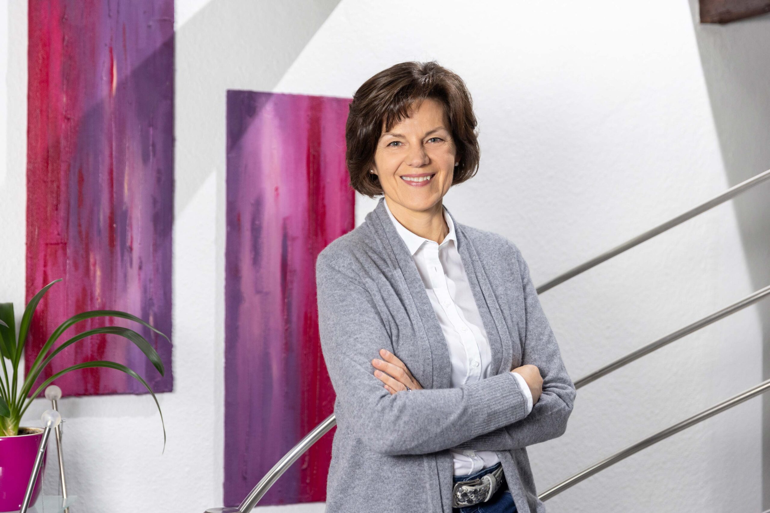 Katrin Pingel professionelle Coachin für Finanzen und Vorsorge aus Unna. Frau Pingel steht im Halbprofil mit verschränkten Armen auf einem Geländer dabei schaut sie freundlich, sympathisch und einladend in die Kamera. Im Hintergrund hängen zwei Gemälde in Rot- und Lilatönen. Das Bild dient zur Illustration der Seite „Was ich anbiete“. Die Themen sind Kompetenzgewinn, Orientierung, Lösungen, Vergütung und Startpaket.