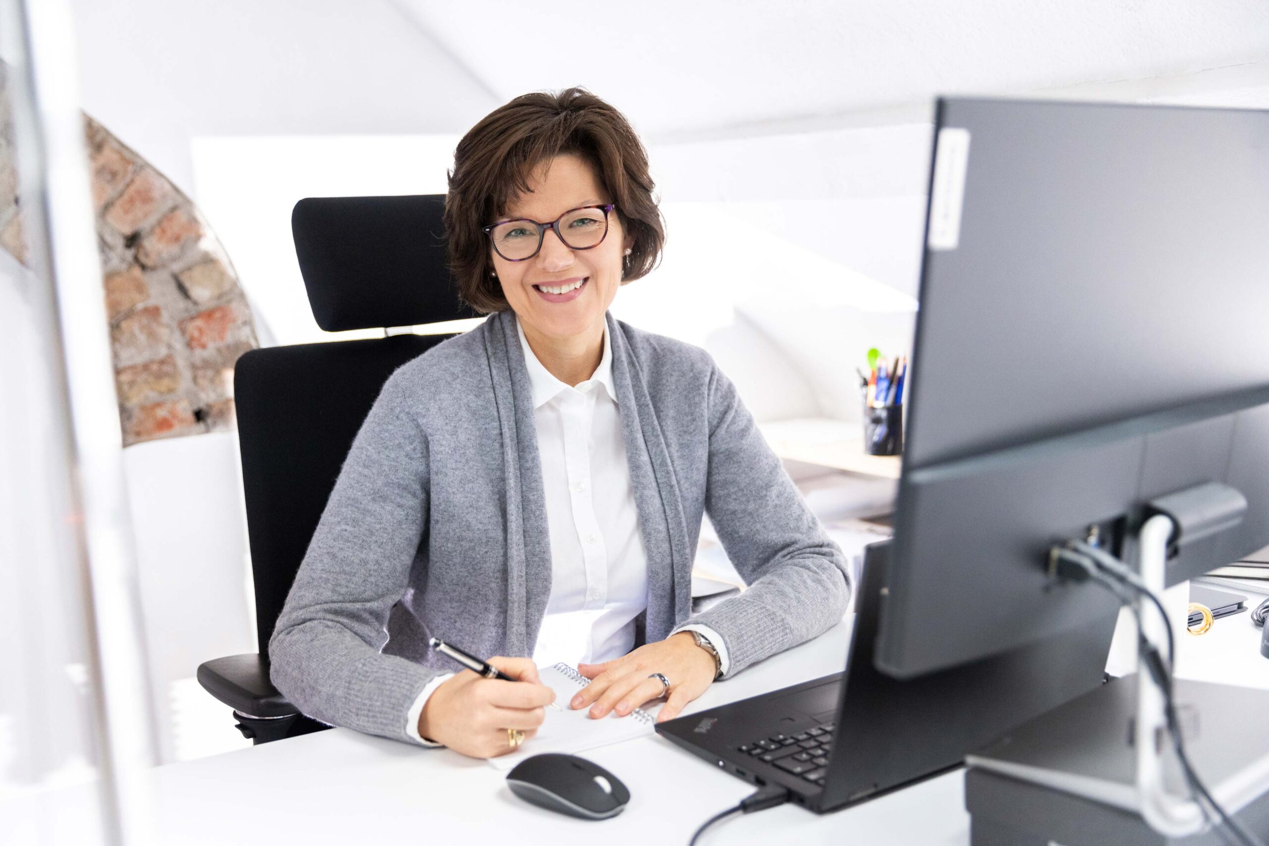 Katrin Pingel professionelle Coachin für Finanzen und Vorsorge aus Unna. Frau Pingel sitzt mit aufgesetzter Brille hinter einem Schreibtisch, ein Stift und Notizblock in den Händen haltend. Sie schaut freundlich, sympathisch und einladend in die Kamera. Im Vordergrund sieht man ein Laptop und einen Monitor. Das Bild wird auf der Seite „Wie ich arbeite“ verwendet. Die Themen sind: präsent, digital, verständlich, methodisch, wirksam, mit dir, mit euch.