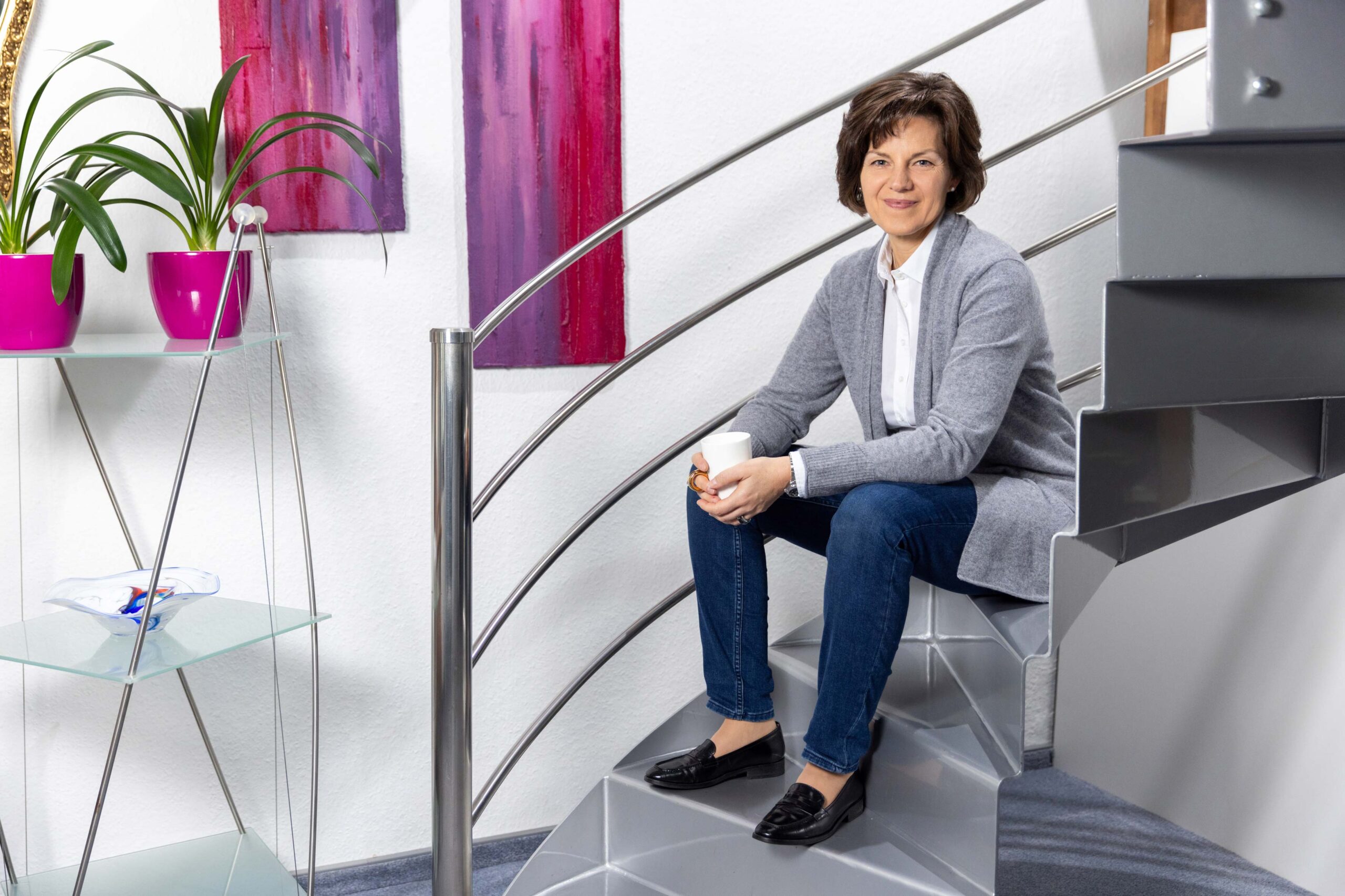 Katrin Pingel professionelle Coachin für Finanzen und Vorsorge aus Unna. Frau Pingel sitzt auf einer Metall-Treppe eine Kaffeetasse in beiden Händen haltend. Die Ellenbogen hat sich bequem auf den Knien liegen. Sie schaut freundlich, lächelnd und sympathisch in die Kamera. Im Hintergrund hängen zwei Gemälde in Rot- und Lilatönen. Das Bild dient zur Illustration der Zitat-Box auf der Startseite zum Thema: „Du allein bist für Deine Entscheidungen verantwortlich – triff‘ Gute!“